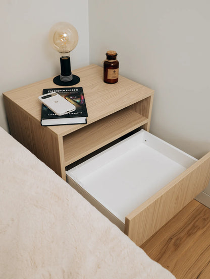 Wooden nightstand with open drawer, books, phone, and lamp on top.