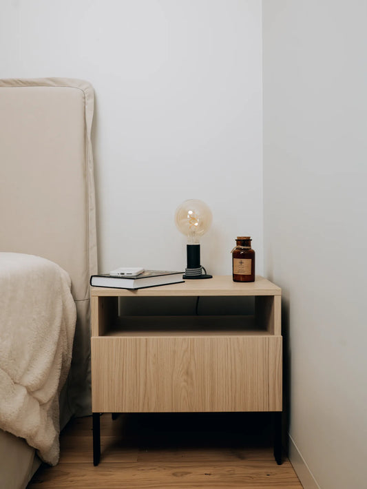 Wooden nightstand with a lamp and book next to a bed in a minimalistic room.